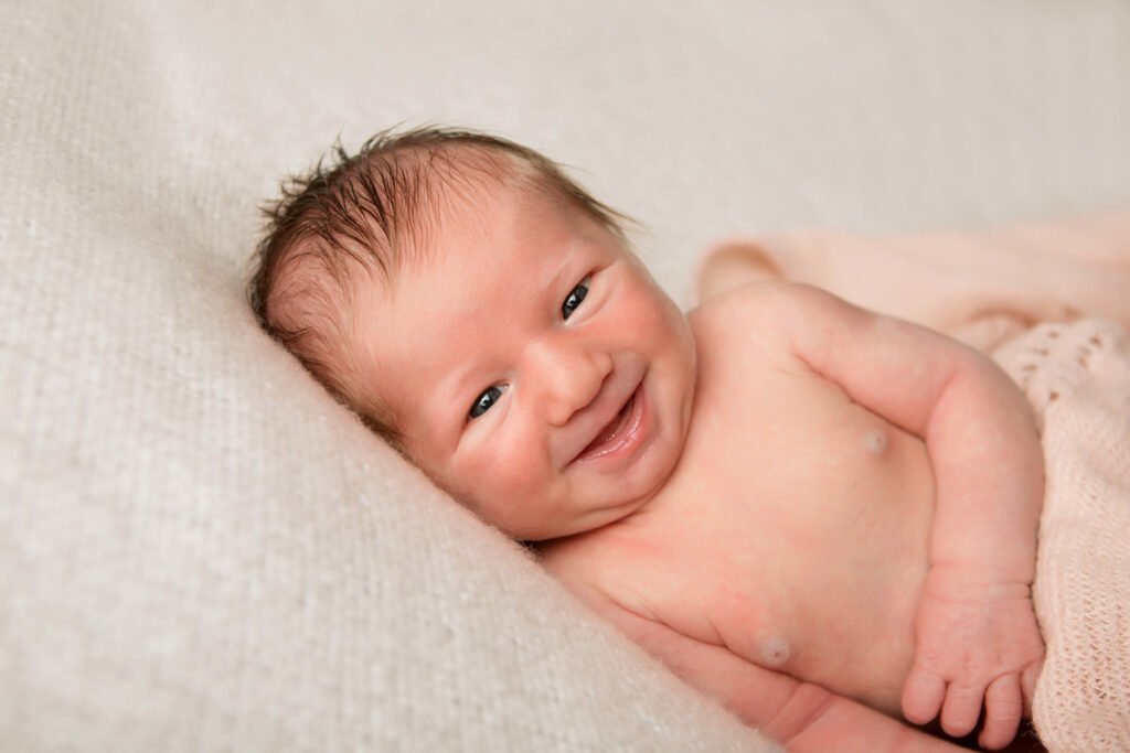 photographe de bébé nouveau-né qui sourit malicieusement allongé sur tissu blanc